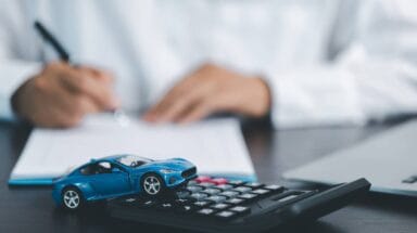 Person signing a document with a car figurine and a calculator in the foreground.